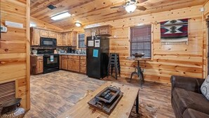 A cabin interior with wooden walls, a kitchen with modern appliances, a dining area with a table and chairs, and a ceiling fan. - Rustic Retreat W/ Hot Tub • Firepit - BBQ (Broken Bow)