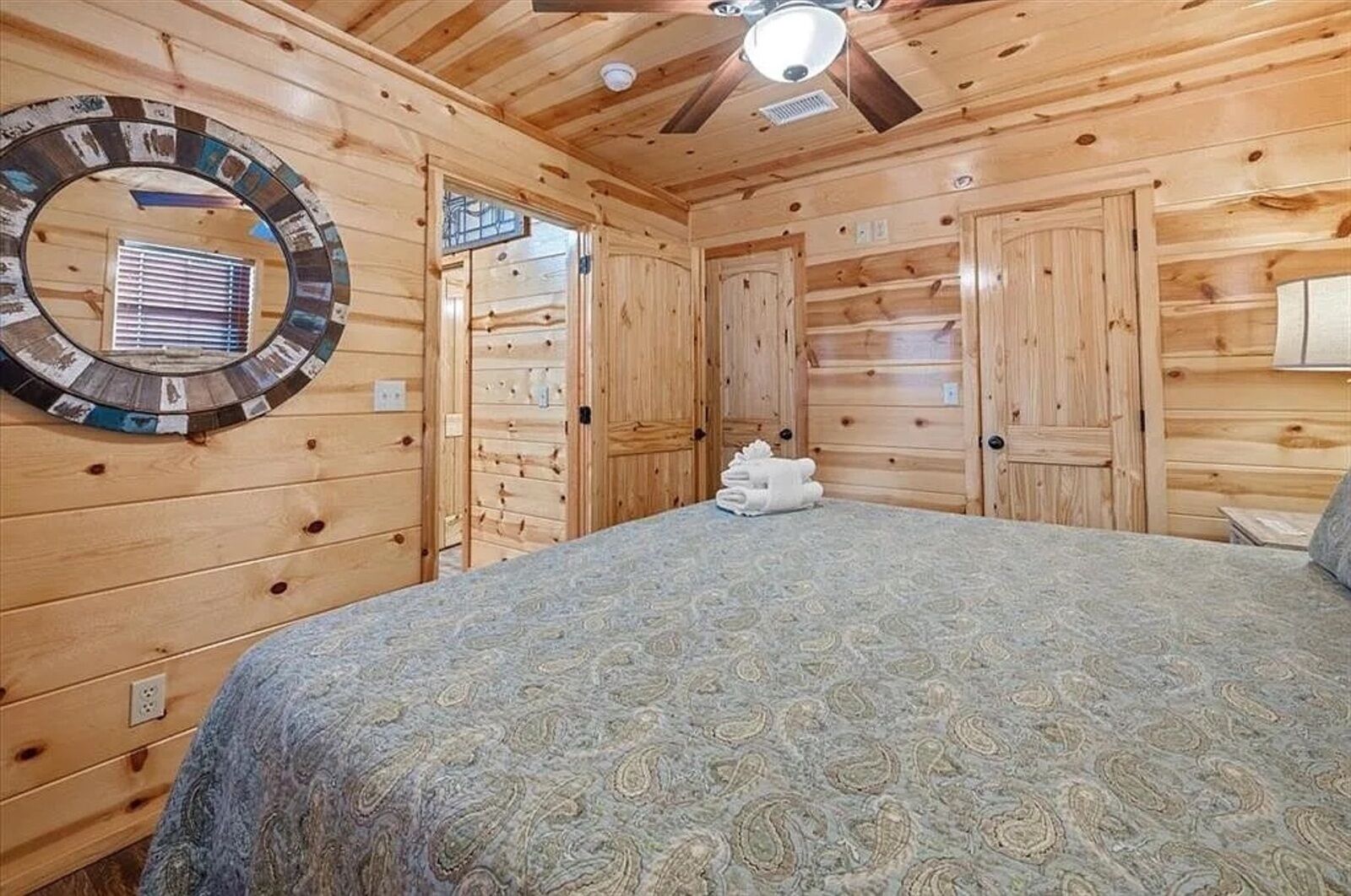 Una habitación tipo cabaña de madera con una cama, un espejo circular y un ventilador de techo.