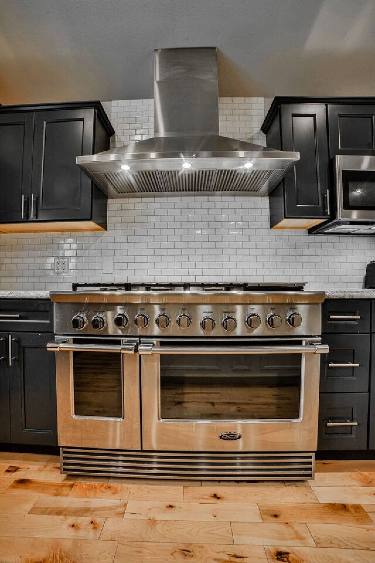 A kitchen with a stainless steel range hood, double ovens, and black cabinets.