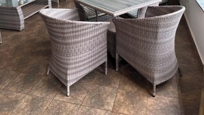 A covered outdoor seating area with wicker furniture and glass-top tables, surrounded by stone pillars and large windows.