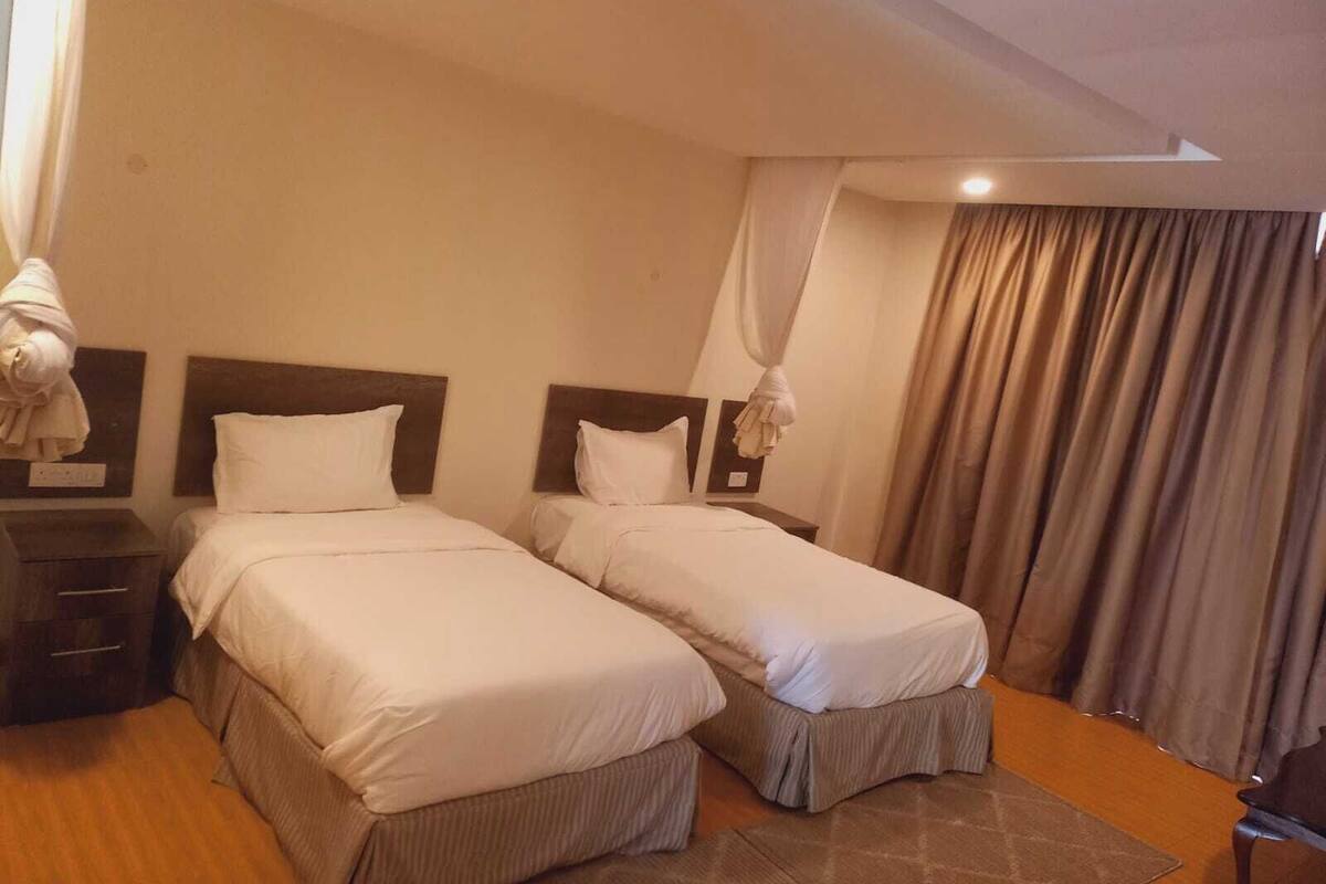 A hotel room with two beds, a nightstand, a lamp, and a curtain.