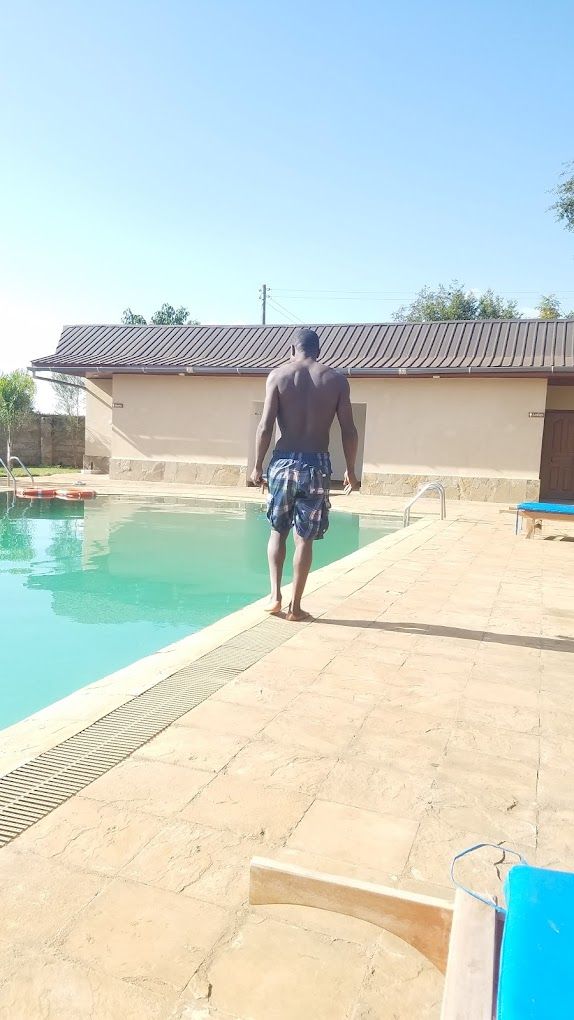 A man walking towards a swimming pool with a tiled deck, a lifeguard stand, and a building with a tiled roof in the background.