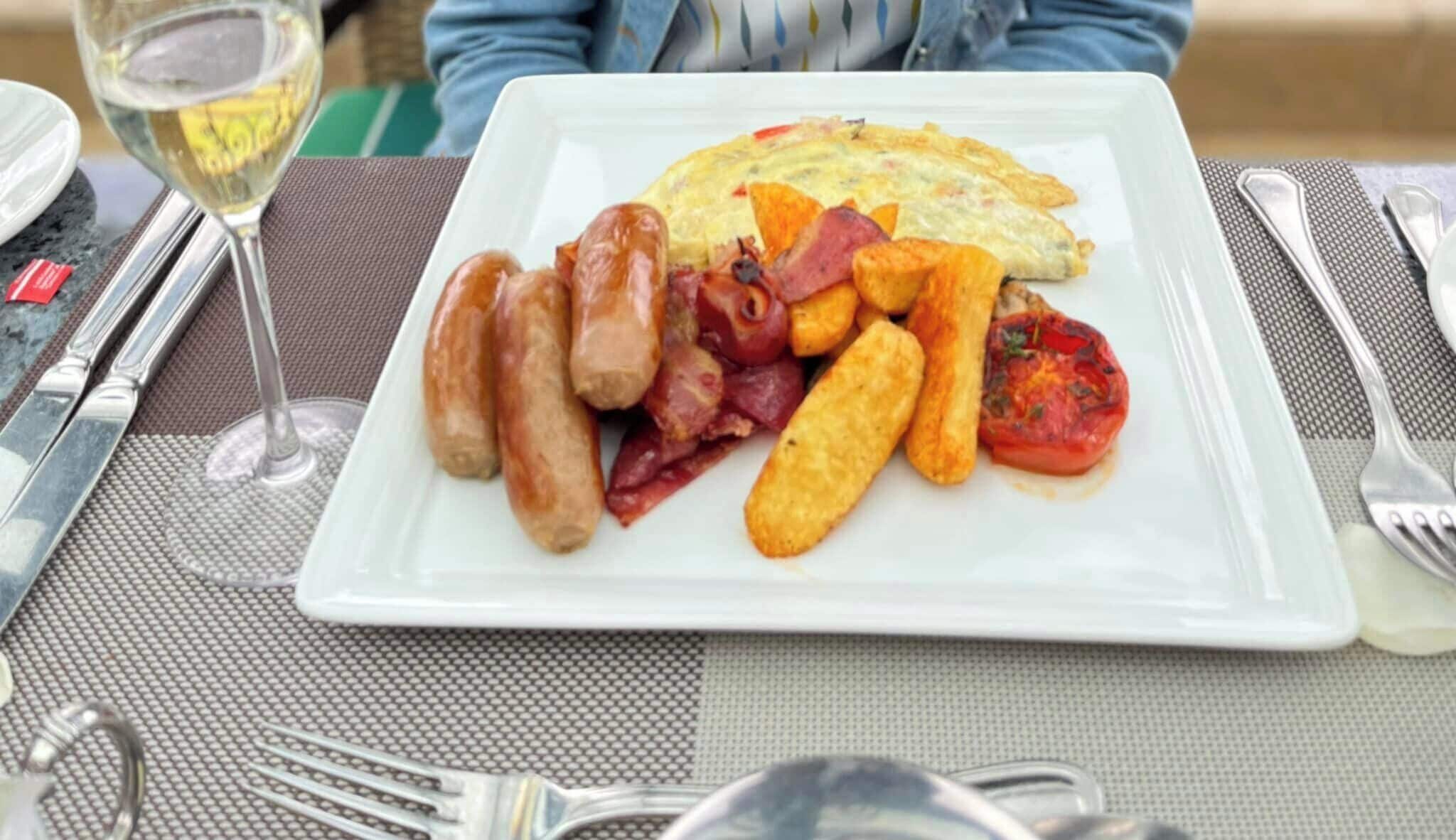 Une assiette de petit-déjeuner comprenant des saucisses, du bacon, des œufs, des pommes de terre rôties et une tomate, le tout servi sur une assiette blanche.