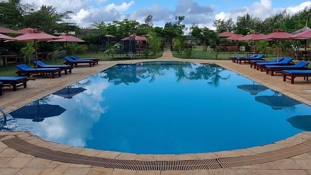 A clear blue swimming pool surrounded by lounge chairs and umbrellas, with a stone pathway leading to a covered area.