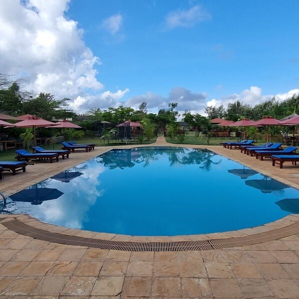 A clear blue swimming pool surrounded by lounge chairs and umbrellas, with a stone pathway leading to a covered area.