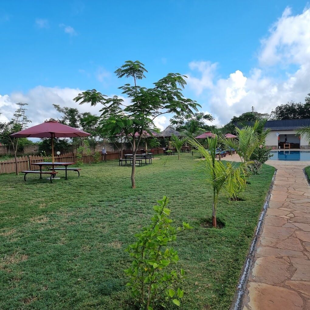 A grassy outdoor area with picnic tables and umbrellas, a small pool, and a building with a blue roof.