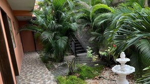 Un sendero estrecho con baldosas estampadas que conduce a una escalera, rodeada de abundante follaje y una fuente para pájaros blanca.