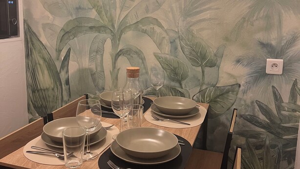 A dining table set with plates, bowls, glasses, and cutlery, with a tropical leaf mural on the wall behind. - Apartment in the heart of Le Creusot (Le Creusot)
