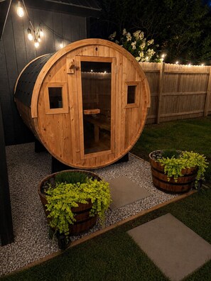A wooden barrel-shaped structure with a door, surrounded by gravel and grass, with two wooden planters with green plants in front.