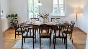 A dining room with a wooden table set for six, surrounded by six chairs. There are two windows with white frames, a hanging light fixture, and a floor lamp. - Idyllic vacation home in the countryside in the Rhön (Oberthulba)