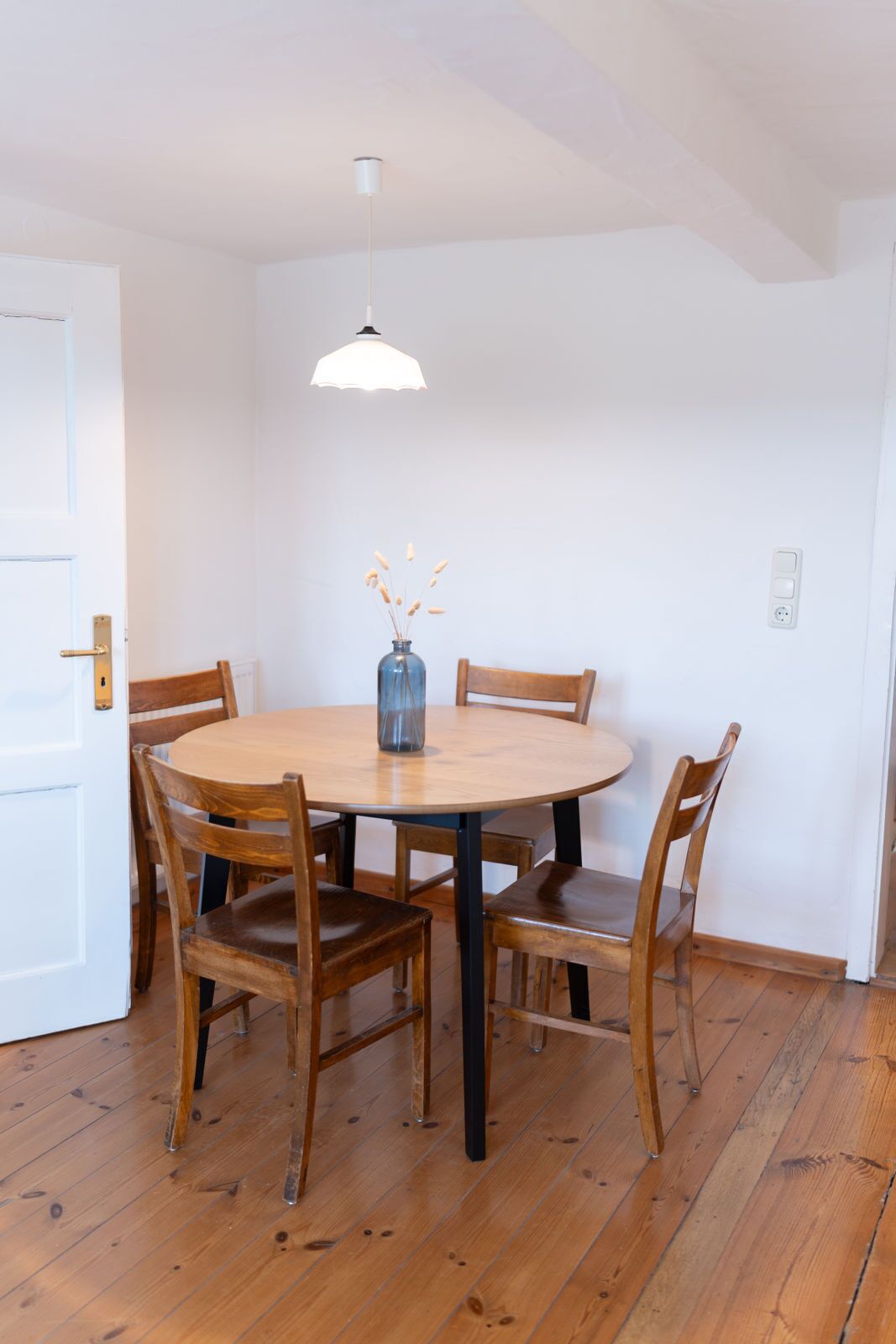 Een eethoek met een ronde houten tafel, vier houten stoelen, een vaas met gedroogde planten, een hanglamp en een witte deur.