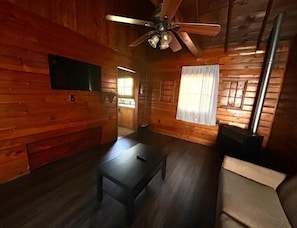 Interior de una cabaña de madera con ventilador de techo, chimenea, televisor montado en la pared y ventana con cortinas.