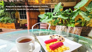 A glass table with a cup of tea and a plate of food, set in a tropical outdoor area with plants and a wooden structure in the background. - Hotel Mayas Blue (Bacalar)