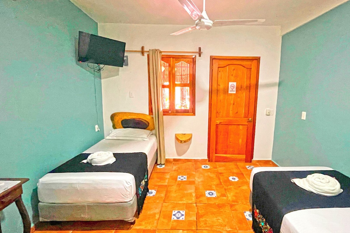 A small, simple room with two beds, a ceiling fan, a TV mounted on the wall, and a door with a sign.
