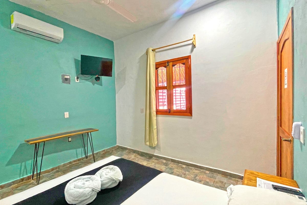 A room with teal walls, a bed, a small table, an air conditioning unit, and a window with a curtain.