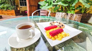 Une table en verre avec une tasse de thé et une assiette de nourriture, placée dans un espace extérieur tropical agrémenté de plantes et d’une structure en bois en arrière-plan.
