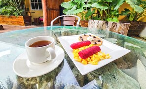 A glass table with a cup of tea and a plate of food, set in a tropical outdoor area with plants and a wooden structure in the background. - Hotel Mayas Blue (Bacalar)