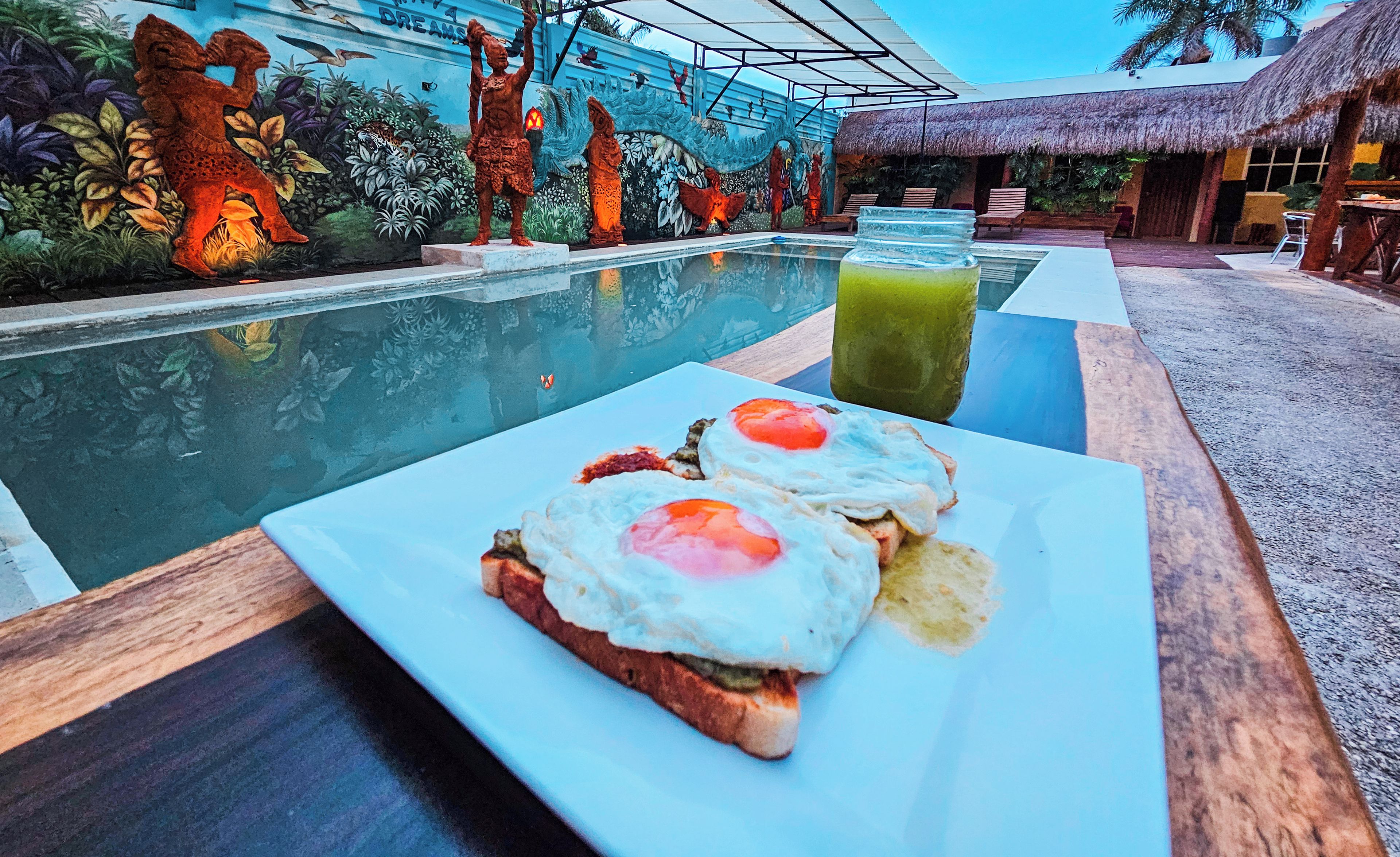 Desayuno con tostadas y huevos sobre una mesa junto a una piscina, con un mural y una estructura de techo de paja al fondo.