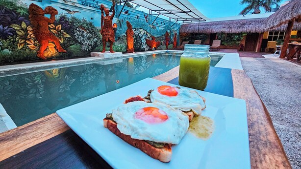 Une table dressée pour le petit-déjeuner, avec des toasts et des œufs, placée près d’une piscine. En arrière-plan, on aperçoit une fresque murale et une structure dotée d’un toit de chaume.
