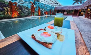 A breakfast setup with toast and eggs on a table by a pool, with a mural and a thatched-roof structure in the background. - Hotel Mayas Blue (Bacalar)