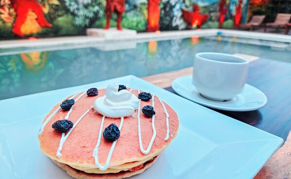A breakfast setting with a plate of pancakes and a cup of coffee, overlooking a pool area with sculptures and a clear sky. - Hotel Mayas Blue (Bacalar)