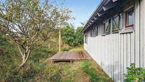 Een houten terras dat leidt naar een wit gebouw met groene luiken, omringd door groen en bomen.