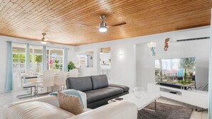 Un salon moderne avec un plafond en bois, un canapé gris, un canapé blanc, un coin repas avec des chaises blanches et un téléviseur fixé au mur.