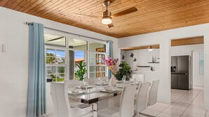 Une salle à manger moderne avec une table en verre, des chaises blanches, un ventilateur de plafond et une vue sur l’extérieur.