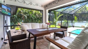 Un patio iluminado por el sol con una mesa de madera, sillas de mimbre y un sofá. Un televisor está montado en la pared, y se ve una piscina a través de las puertas de vidrio.