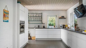 Une cuisine moderne avec des meubles blancs, un four encastré, une fenêtre donnant sur un espace verdoyant et une étagère avec des assiettes.