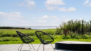 Deux chaises au design minimaliste, avec une structure en fil de fer noir, sont placées sur une terrasse carrelée qui donne sur un champ verdoyant et un plan d’eau.