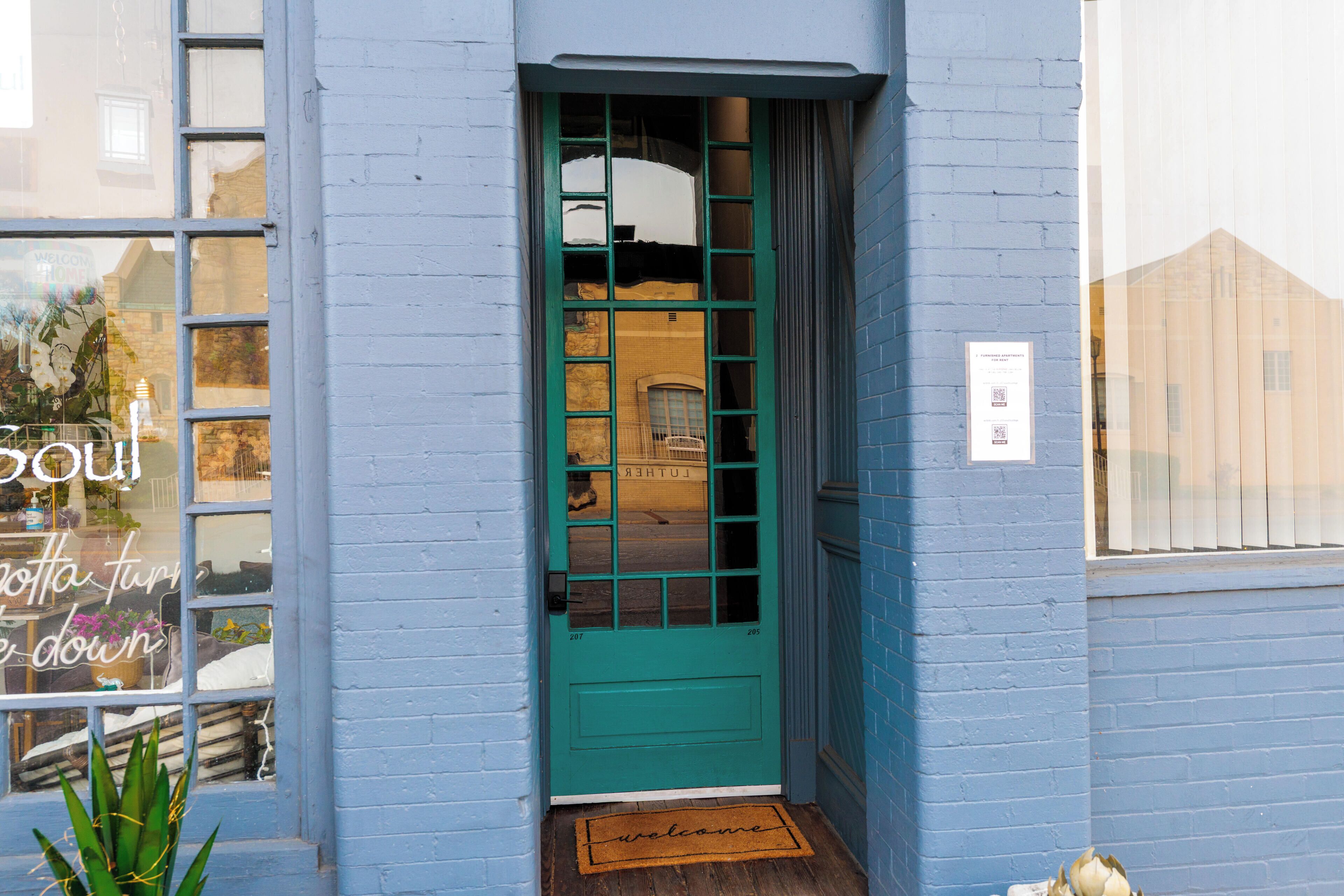 A teal door with a window above it, a welcome mat at the entrance, and a sign on the wall.