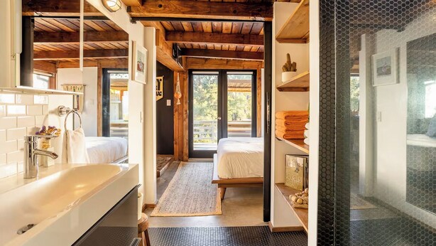 Ein modernes Badezimmer mit einem großen Waschbecken, einem Spiegel und einer Dusche. Eine Holzbalkendecke und ein Blick ins Freie durch eine Glastür.