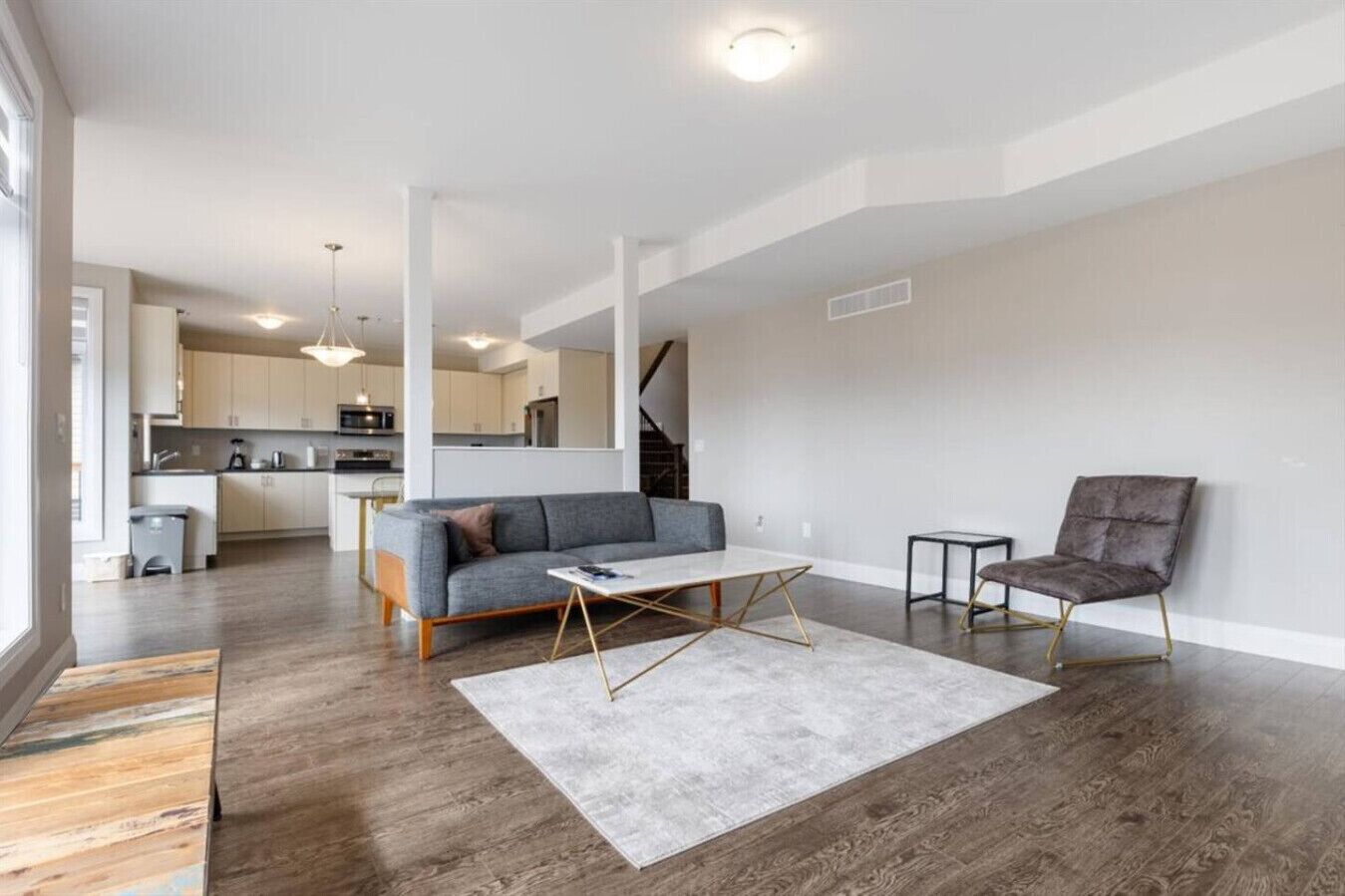 A modern living room with a grey sofa, a coffee table, and a chair. There is a kitchen in the background with white cabinets and a staircase.