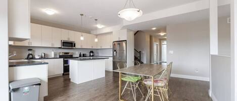 A modern kitchen with white cabinets, a central island, stainless steel appliances, and a dining area with a wooden table and chairs.