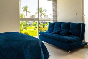 A room with a large window, a blue sofa, and a bed with a blue cover.