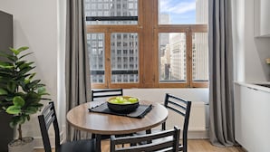 Comedor con una mesa redonda, sillas, un bol de manzanas, una planta en maceta y vistas de edificios urbanos a través de un amplio ventanal con cortinas.