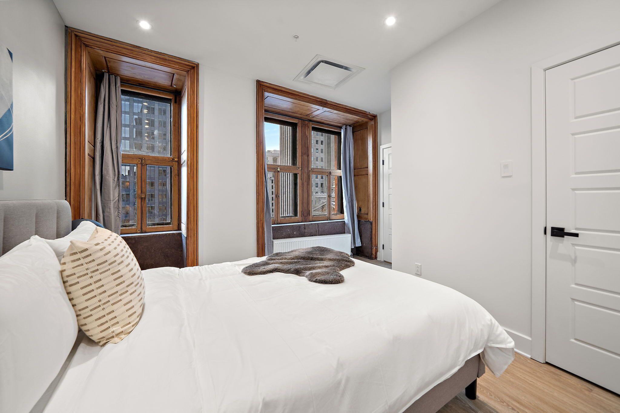 Quarto com uma cama grande, duas janelas com cortinas, um radiador e uma porta branca.