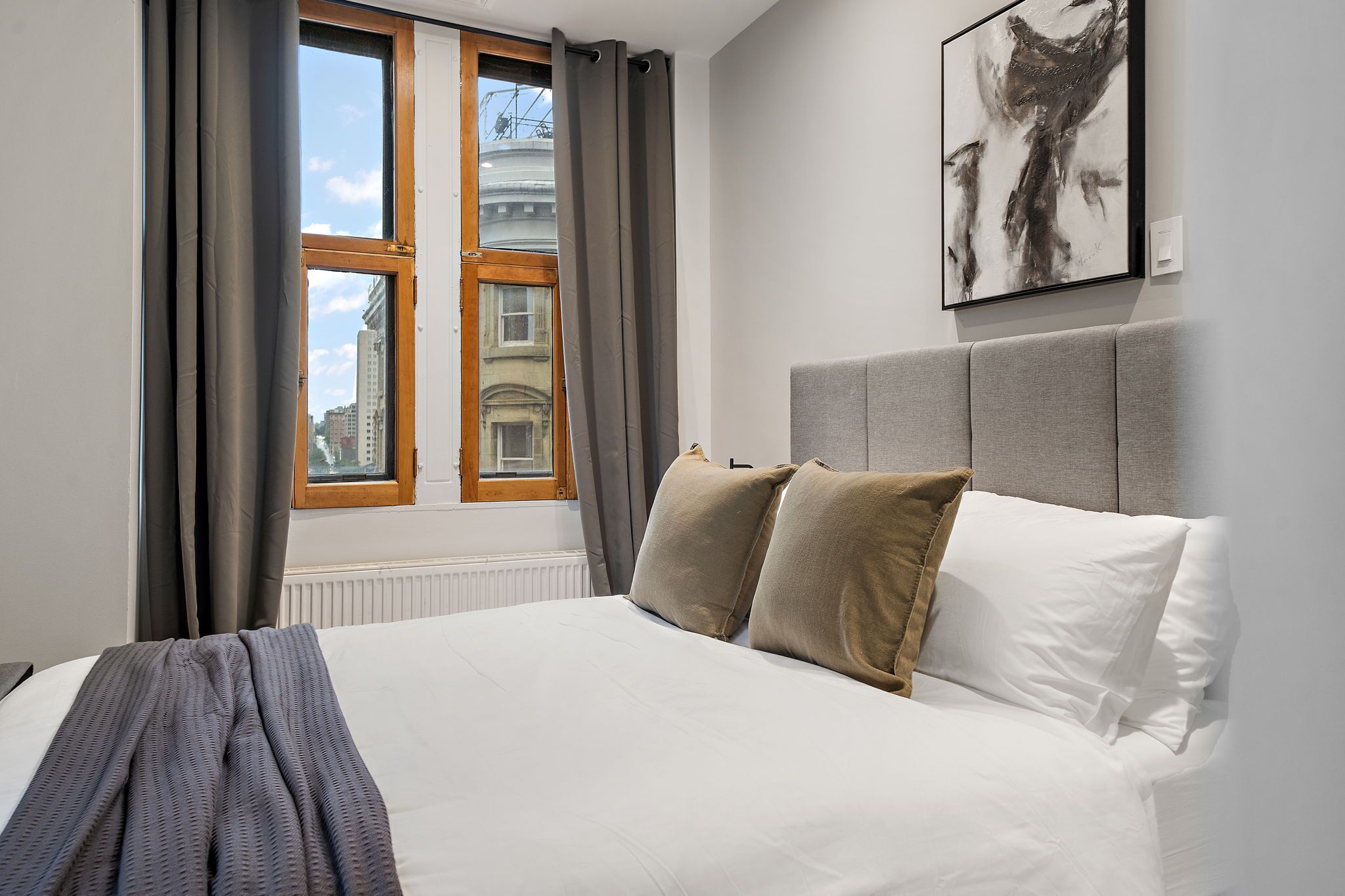 Un dormitorio con una cama grande, cortinas grises, una ventana con vistas a edificios y un cuadro abstracto enmarcado en la pared.