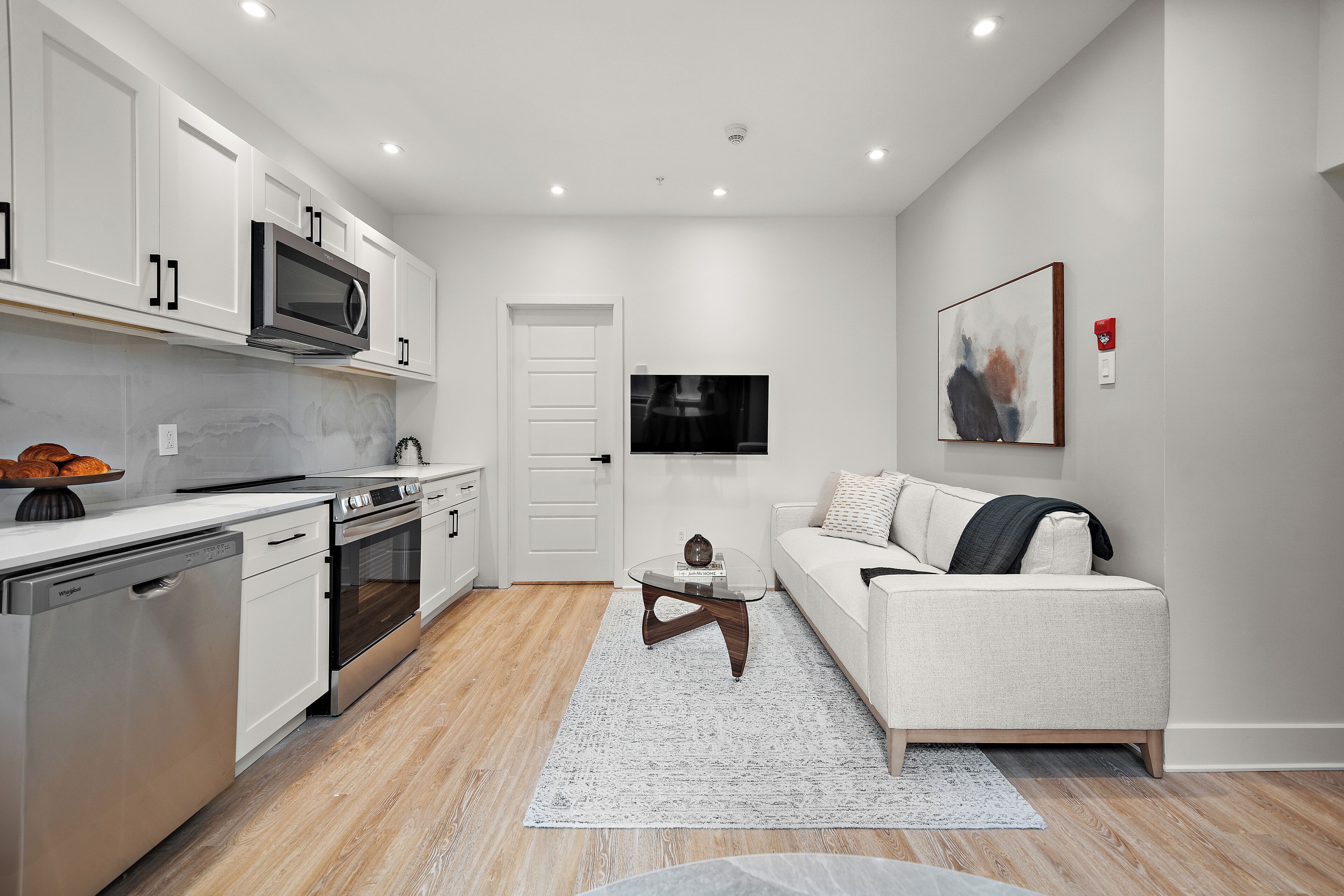 A modern kitchen with stainless steel appliances, a dining area with a glass table, and a living room with a sofa and a painting on the wall.