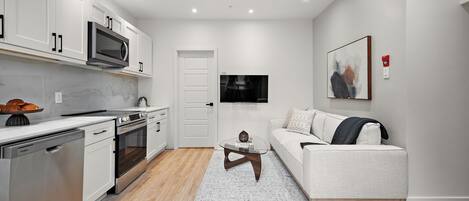 A modern kitchen with stainless steel appliances, a dining area with a glass table, and a living room with a sofa and a painting on the wall.