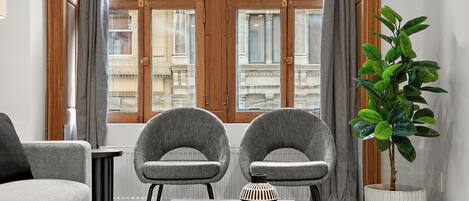 A modern living room with two grey armchairs, a wooden coffee table with a magazine, a potted plant, and a view of a building through large windows.