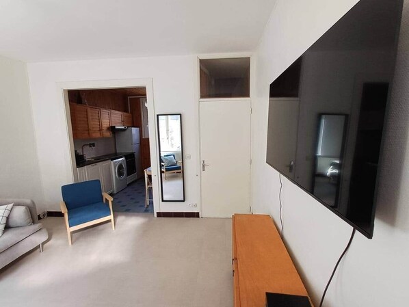 Un salon avec un canapé, un fauteuil bleu, un téléviseur et un banc en bois. Une porte donne accès à une cuisine équipée d’un lave-linge.