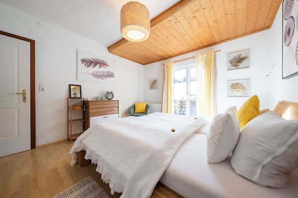 A bedroom with a wooden ceiling, a bed with white bedding, a bedside table, a chair, and a window with curtains.