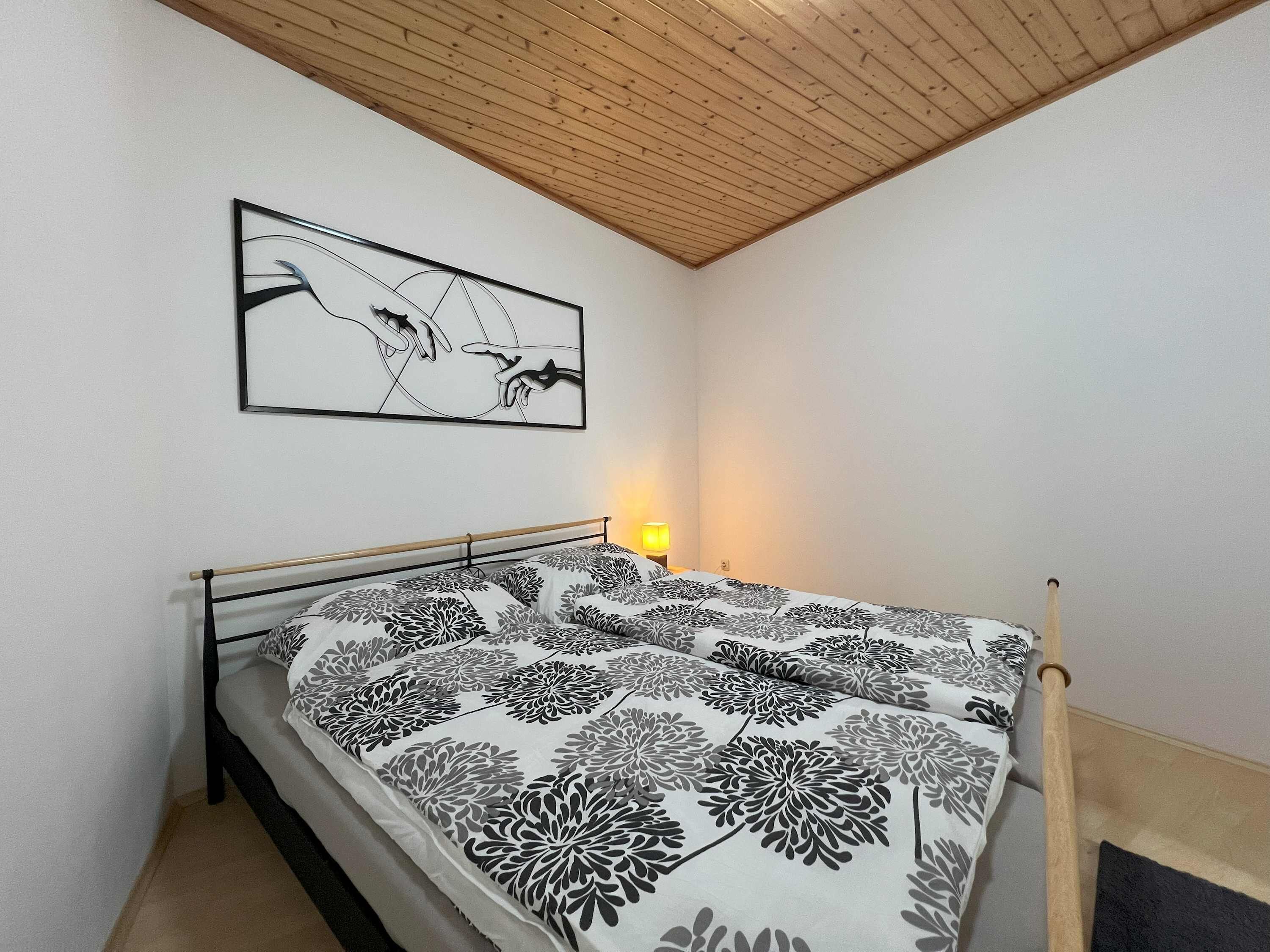 A bedroom with a bed, a wooden ceiling, a wall-mounted artwork, and a bedside lamp.