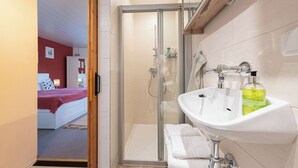 A modern bathroom with a glass shower enclosure, a white sink with a green bottle, and a bed visible through an open door. - Krokus Studio | Relax & Natur | Parking | Bikes (Altaussee)