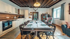A dining area with a wooden table set for four, surrounded by chairs, in a room with wooden beams on the ceiling and a large window with curtains. - Strauss Suite | Kitchen | Parking | Garden (Altaussee)