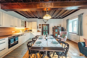 A dining area with a wooden table set for four, surrounded by chairs. A kitchen is visible to the left with white cabinets and a stove.