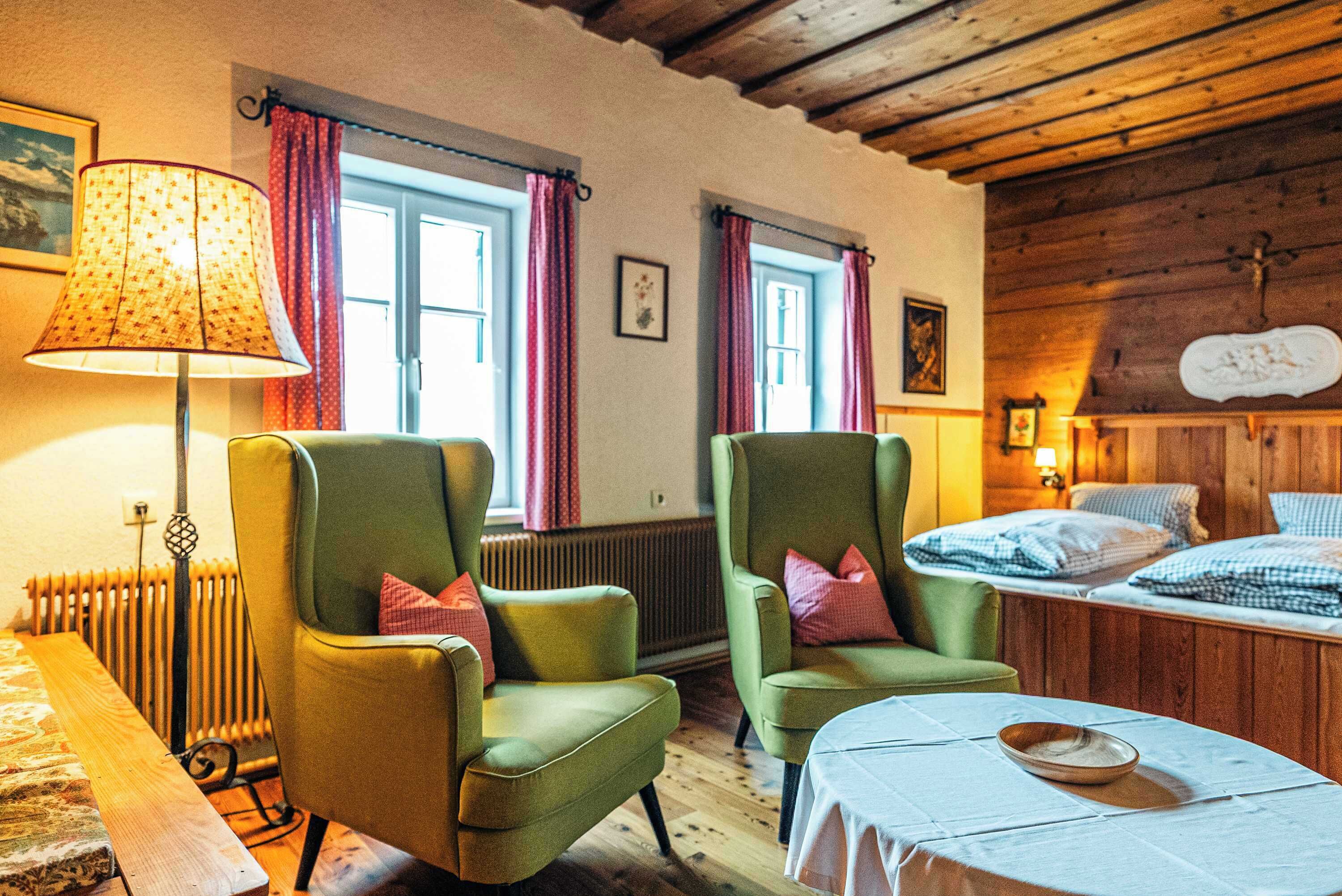 Ein Zimmer mit zwei grünen Sesseln, einem Holztisch, einem Bett mit karierter Bettwäsche, einem Heizkörper und einem Fenster mit Vorhängen.