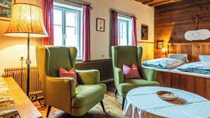 Ein Zimmer mit zwei grünen Sesseln, einem Holztisch, einem Bett mit karierter Bettwäsche, einem Heizkörper und einem Fenster mit Vorhängen.